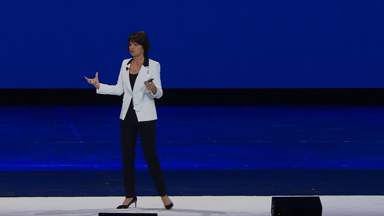 Motivational Speaker Lisa McCarthy is delivering a keynote speech for an audience from the stage. She is wearing a white blazer and holding a clicker